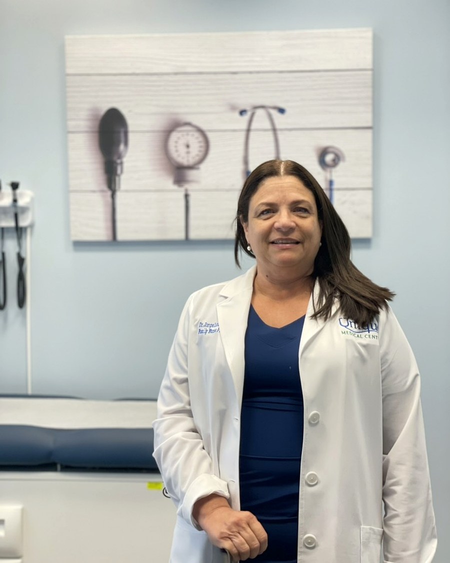 Dr. Jorgelina D'Fana Valdes, APRN, smiling in her exam room wearing a white coat over navy scrubs